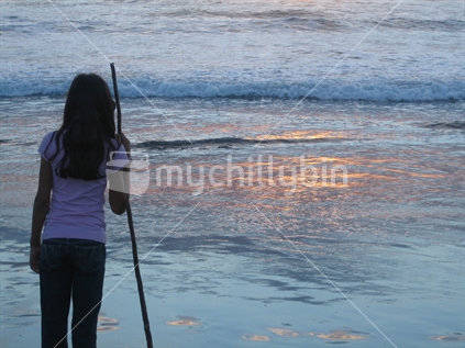 Young girl holding stick contemplating the sunset at Bethells Beach
