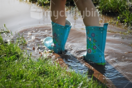 A child wearing gumboots splashes in a muddy puddle (selective focus)