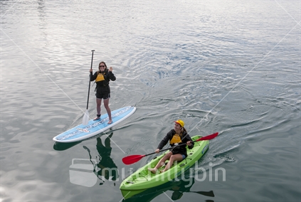2 young women having fun with water sport activities. One is on a paddle board and is waving to the photographer. The other is paddling a canoe