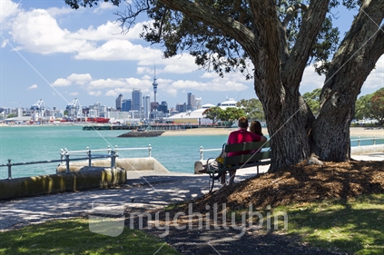 Couple sitting under a tree on Devonport seafront with Auckland city CBD in the background.