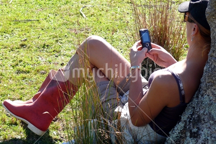 Girl in gumboots resting against a cabbage tree on rural property, texting