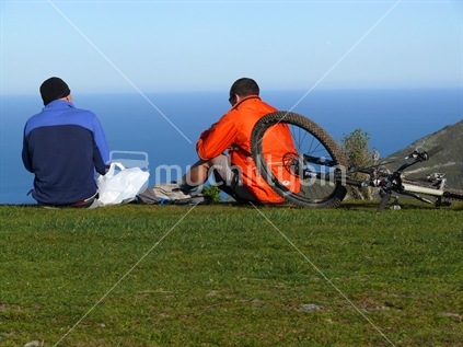 Two mountain bikers resting on green hill overlooking sea with lunch