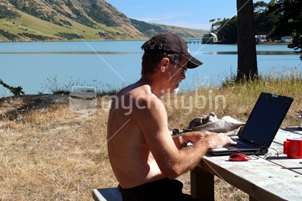 Man on holiday working on laptop on picnic bench by the New Zealand coast