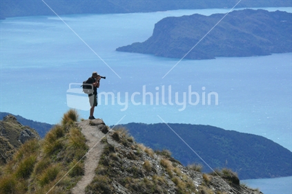Man with telephoto lens taking photo of Lake Wanaka from Roy Peak with track in foregrpound