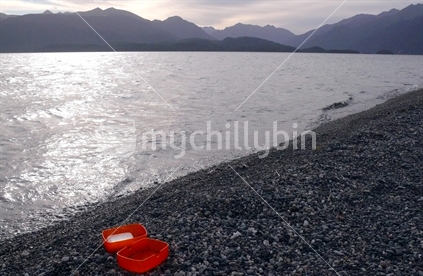 An orange plastic soap box with soap lying on pebbled beach beside pristine lake in sunset