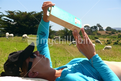 Young woman lying in sun reading book with sheep in background.