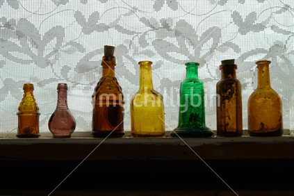 A line of old bottles on a wooden shelf with lace curtain