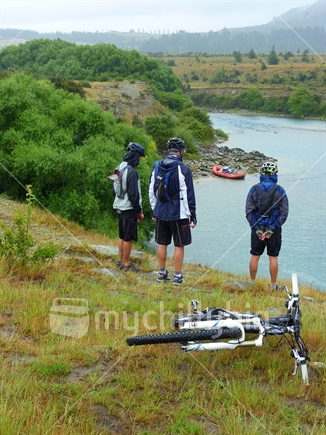 Three mountain bikers resting whilst looking over the Clutha River and a raft. 
