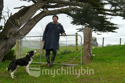 Farmer wearing swandri leaning on farm gate with sheep dog by his side