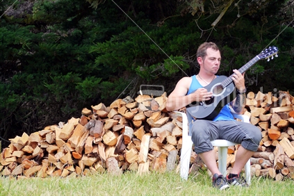 A young man playing a guitar with stacked firewood behind