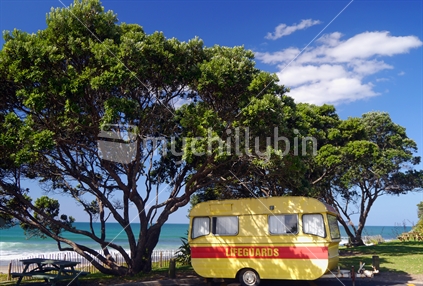 Old lifesaving caravan, parked under pohutukawa trees and blue sky.