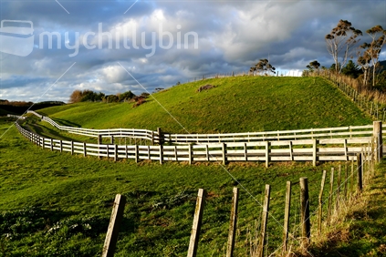 Wooden fence running across a hilly paddock, with a fenced driveway leading into the distance
