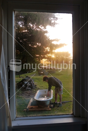 Looking through dirty window at man testing the heat of water in outdoor bath,