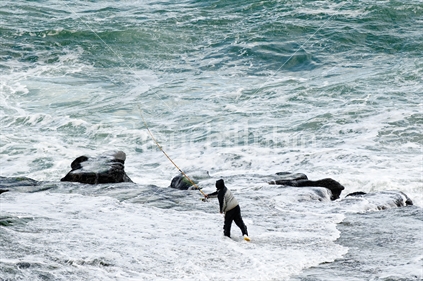 Lone fishermen fishing from wave swept rocks at Muriwai surf beach