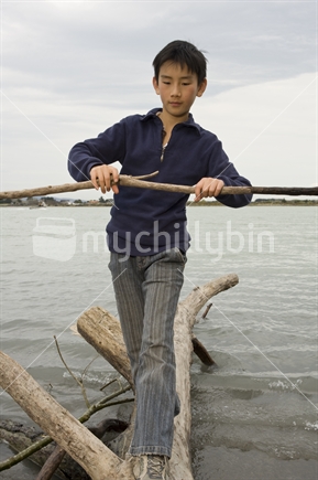 11 year old Asian boy balancing on log in water near Kairaki / mouth of Waimakariri River 