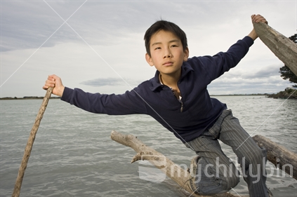 11 year old Asian boy balancing on log in water near Kairaki / mouth of Waimakariri River