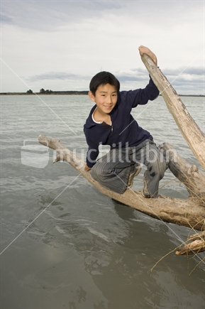 11 year old Asian boy balancing on log in water near Kairaki / mouth of Waimakariri River