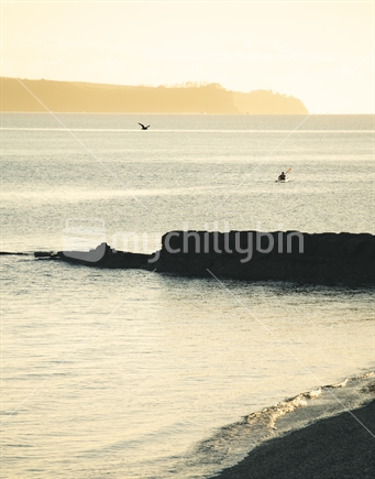 Shoreline, rick (focus) and sunrise kayaker, Auckland
