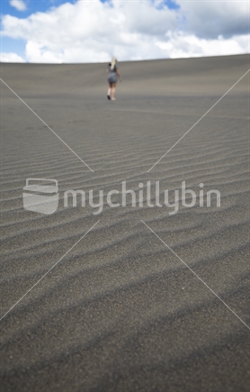Focus on black sand dunes with female walking in distance, Lake Waimanu, Bethells beach, Auckland