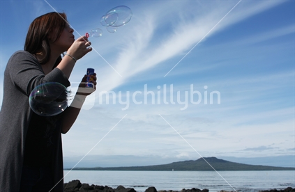 woman blowing soap bubbles with Rangitoto Island in the background, Takapuna, North Shore, Auckland, New Zealand
