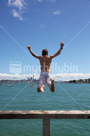 A boy jumping off Devonport wharf, North Shore, with Auckland CBD and Skytower in the background.
