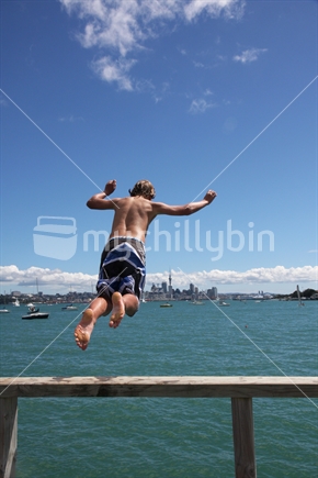 A boy jumping off the Devonport wharf, North Shore, with Auckland CBD and Skytower in the background.

