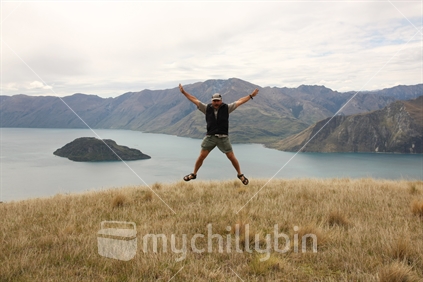 Man jumping in the air on top of the mountain with breathtaking views of Lake Wanaka