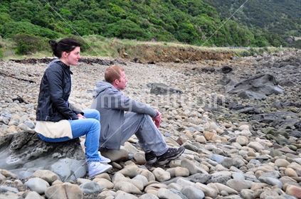 Couple sitting on rocky beach, on New Zealand coastline. 