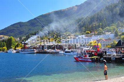 Man in swimming togs on waterfront in Queenstown, wiht smoke from the historic Earnslaw drifting across. 
