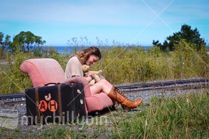 Young woman with teddy bear and suitcase sitting in an easy chair beside railway line on the Kaikoura coast