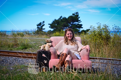 Young woman with teddy bear and suitcase sitting in chair beside railway line on the Kaikoura coast
