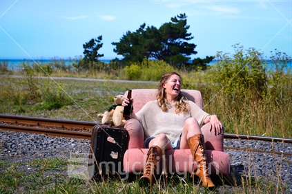 Bear and Beer; young woman with teddy bear and suitcase sitting in chair beside railway line on the Kaikoura coast 