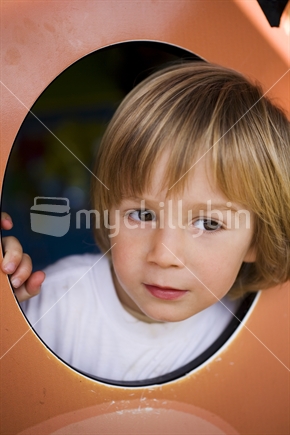 A young blonde boy peers out of a cutout oval at a kids playground
