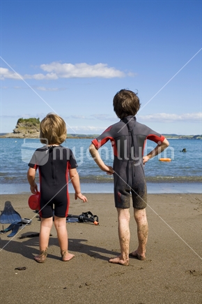 Two boys in wetsuits from behind looking out over the ocean at Mathesons Bay, Northland
