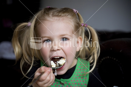 A blonde girl with pigtails eating a large spoon of ice cream