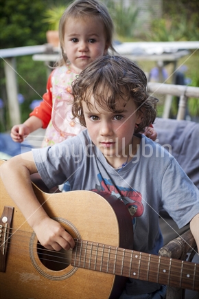 A young boy playing guitar while his sister looks on