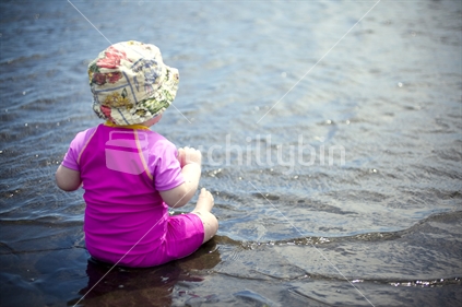 Baby girl in pink, siting at waters edge at Piha, Auckland, New Zealand.