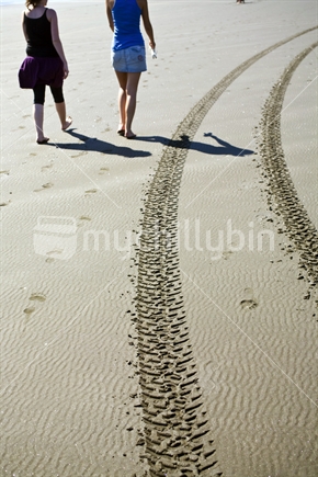 Two girls walking along the beach next to tire marks
