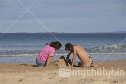 Teenaged siblings sit together and build sandcastles on the beach. 