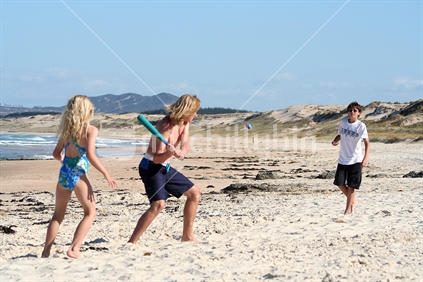 Three youngsters play a game of bat and ball at a New Zealand beach in summer. 