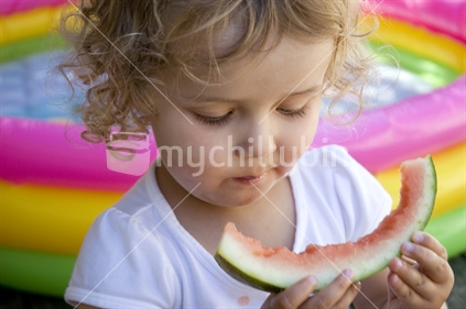 Young girl eats a watermelon - beside a paddling pool on a hot summer's day