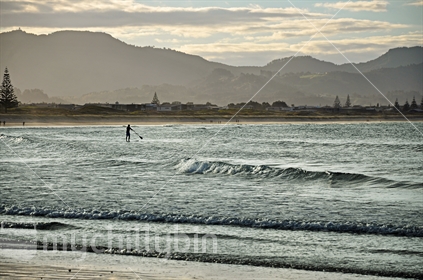 A paddleboarder at dusk on Omaha beach, Rodney District, North Island, New Zealand.
