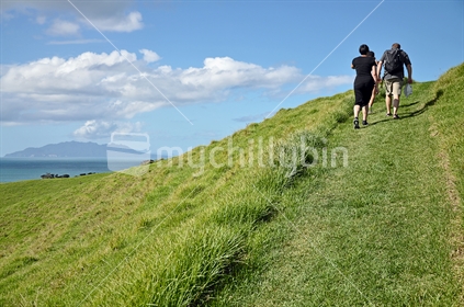 Family climb a grassy hill, Little Barrier in the distance