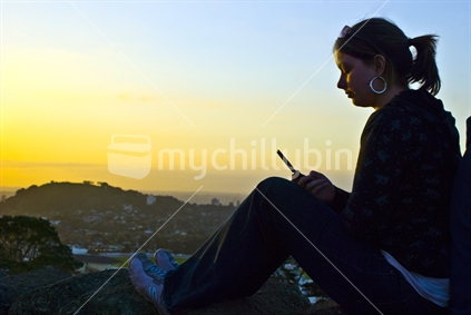 Young lady texting on mobile phone in sunset overlooking Auckland city
