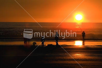 A Family playing and fishing at the beach in front of a setting sun, New Zealand