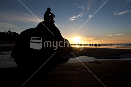 A man sitting on a large rock at a beach watching the sun set, New Zealand