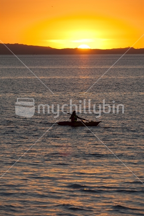 A kayaker (focus) paddling in front of a setting sun.