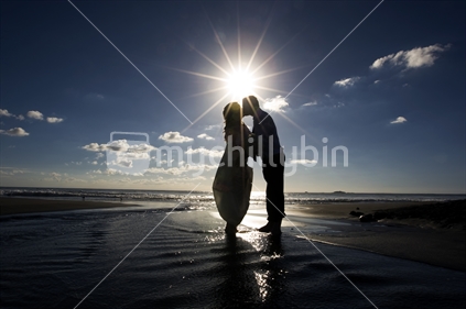 A wedding couple kiss and embrace at a New Zealand beach, in front of a setting sun.