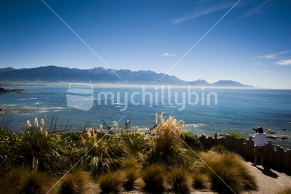 A tourist taking a photo of the Kaikoura coastline and Kaikoura ranges from a walkway lookout point.