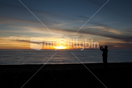 A tourist taking photos of a sunrise on the beach at Kaikoura, East Coast of the South Island, New Zealand.